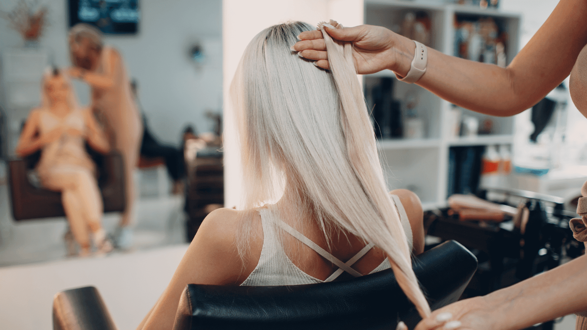Blonde woman getting her long hair styled at a salon, reflected in a background mirror.