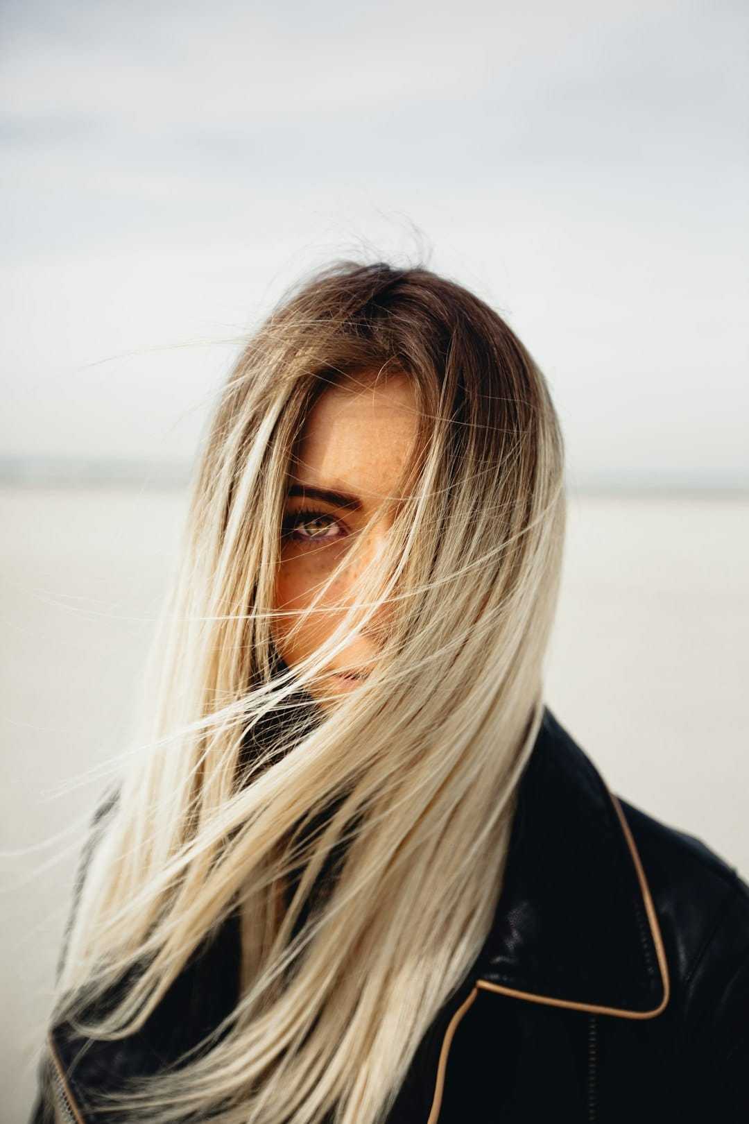 Woman with long hair blowing across her face, wearing a black leather jacket on a beach.