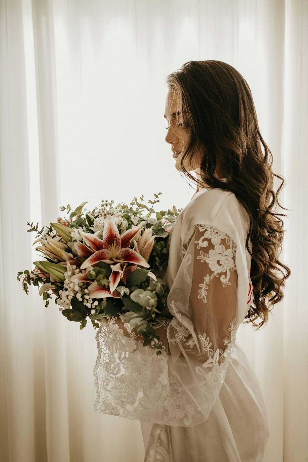Bride in lace robe holding a bouquet with lilies, standing by a softly lit window.