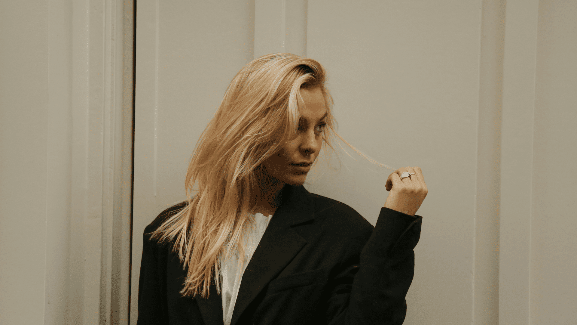 Blonde woman in a black blazer looks sideways against a neutral wall.