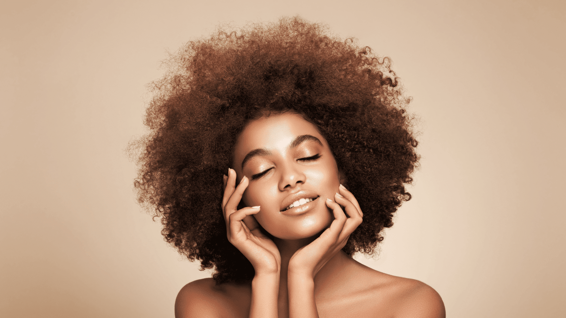 Woman with curly hair smiling with closed eyes against a neutral background.