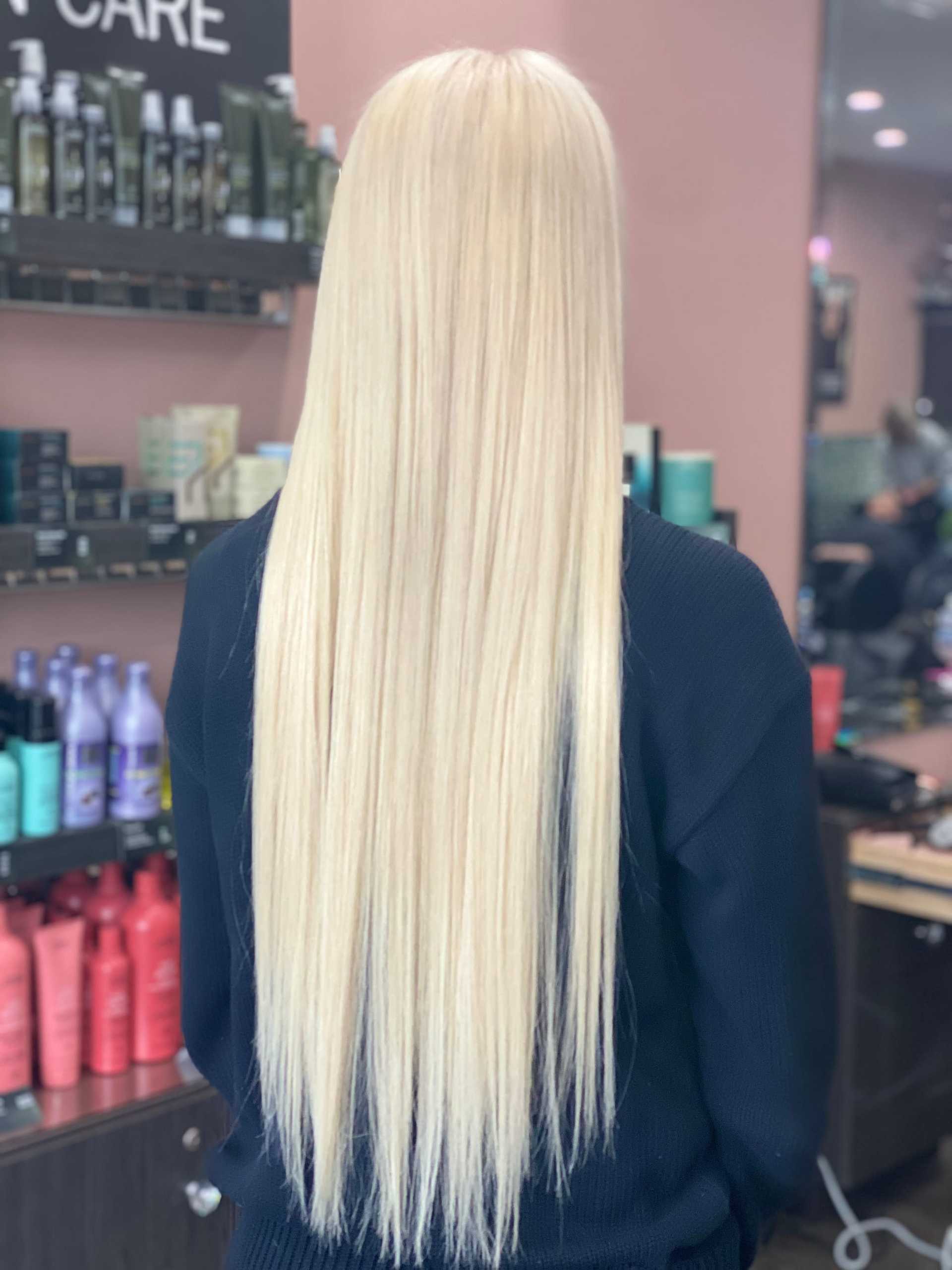 Blonde-haired person with long straight hair inside a salon with products on shelves.